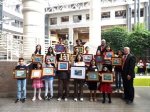 The 2025 Art Calendar Contest, hosted by Public Works, showcases a vibrant celebration of youth creativity revolving around the theme "Reimagine Water." This image captures the essence of the event, featuring a diverse group of students proudly displaying their artworks. Each child holds a framed piece that exemplifies their unique artistic interpretation of water's significance in our lives, embodying the contest's goal to raise awareness about natural water resources and their management. The excitement and sense of accomplishment are palpable, highlighting the importance of engaging young minds in environmental consciousness. The backdrop of the event emphasizes a modern architectural setting, suggesting a physical space that is both open and welcoming. Large glass windows and lush greenery inside provide a refreshing atmosphere, symbolizing the very theme of water and nature that the artworks represent. Standing amidst towering columns, the students form a cohesive group, underscoring their collective achievement and camaraderie. This visually striking environment not only enhances the students' artistic expressions but also aligns with the contest's mission of fostering community connection and environmental stewardship. The array of artworks seen in the students' hands spans various styles and techniques, showcasing a rich tapestry of perspectives on water. From vibrant, energetic depictions of flowing rivers to more serene interpretations of still lakes and ocean waves, the pieces reflect an understanding of water's multifaceted role in our ecosystem. Each artwork serves as a personal narrative, allowing viewers to glimpse the artists' individual relationships with water — whether through play, conservation, or reflection. The contest challenges participants to not only express their creativity but also to explore deeper themes of sustainability and responsible water management. The presence of community leaders and representatives in the background underscores the importance of support for youth initiatives in promoting environmental awareness. Their engagement with the students highlights a collaborative effort to instill values of stewardship and responsibility regarding natural resources. This event not only recognizes artistic talent but also reinforces the imperative of nurturing a future generation that is informed, inspired, and capable of tackling pressing environmental challenges. The triumph of the 2025 Art Calendar Contest serves as an encouraging reminder of the transformative power of art in bridging community efforts and fostering a deeper appreciation for our most vital resource: water.